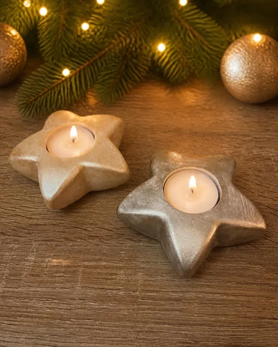 Duas estrelas porta-vela, uma dourada e outra prateada, com velas de chá acesas, sobre madeira, entre ramos de pinheiro e bolas douradas.
